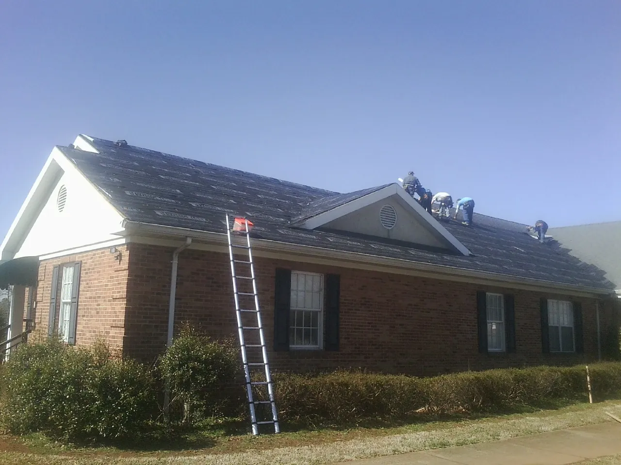 Crew tearing shingles off a brick ranch roof with a ladder set — Metal Roof Repair work by our crew in Lacona