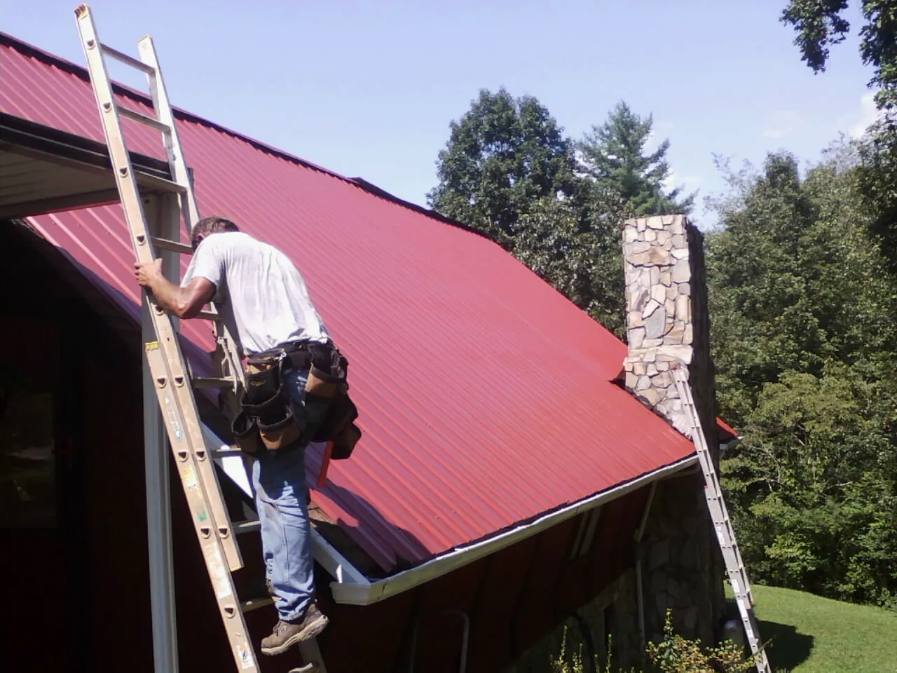 Roofer on a ladder beside a red metal roof and stone chimney — Soffit & Fascia Repair by Roofing Brothers in Lacona